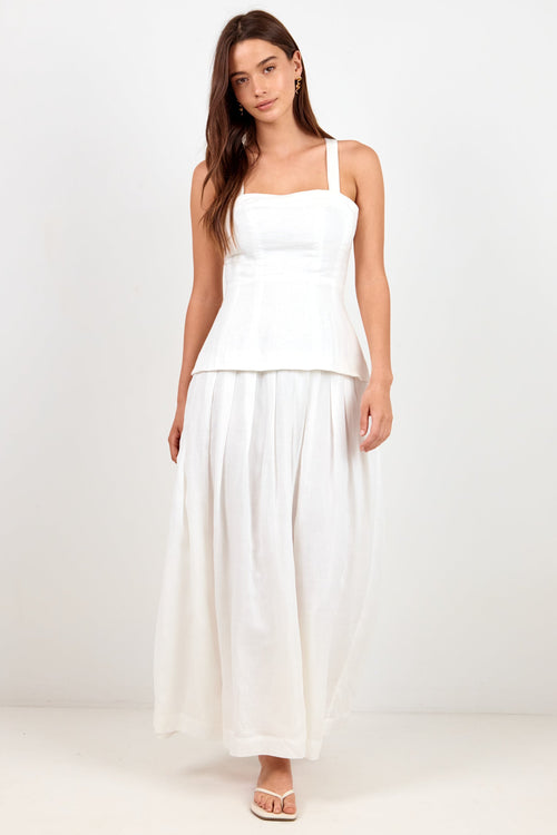 A woman modeling a white sleeveless maxi dress with a peplum waist and flowing skirt, standing against a plain white studio background.
