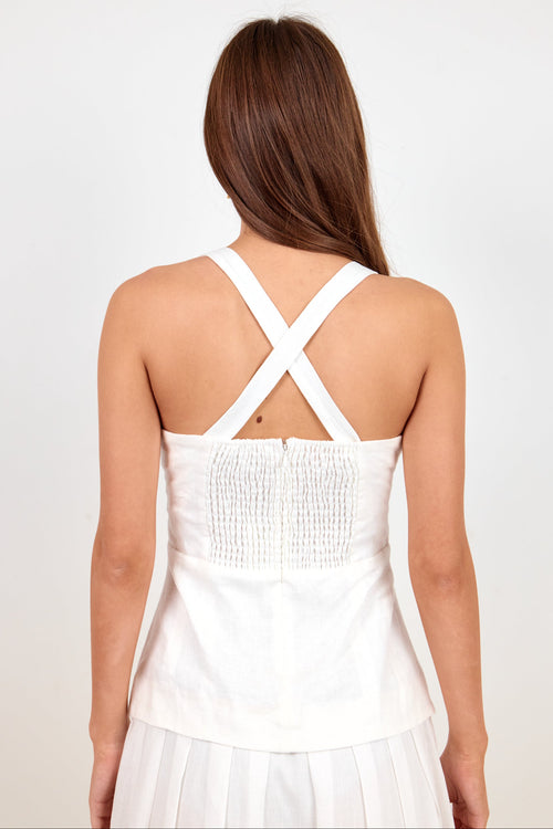 A woman viewed from the back wearing a white sleeveless top with crisscross shoulder straps and a smocked bodice, paired with a white skirt, against a plain light background.