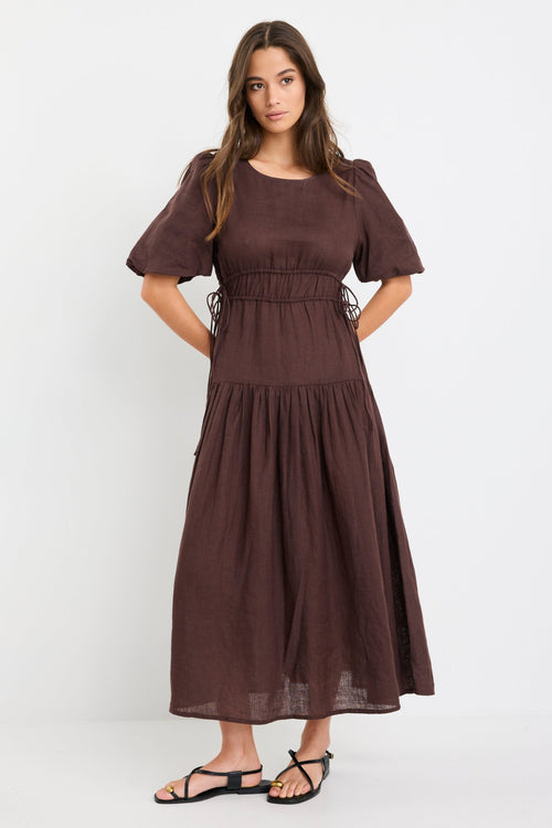 Woman wearing a chocolate brown linen midi dress with puff sleeves, tie waist detail, and tiered skirt, styled with black strappy sandals.