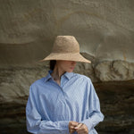 A woman wearing a wide-brimmed straw hat and a light blue striped button-up shirt stands in front of a large natural rock formation.