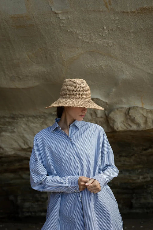 A woman wearing a wide-brimmed straw hat and a light blue striped button-up shirt stands in front of a large natural rock formation.