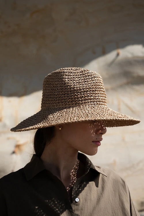 A woman in profile wearing a wide-brimmed woven straw hat in a warm sandy environment.