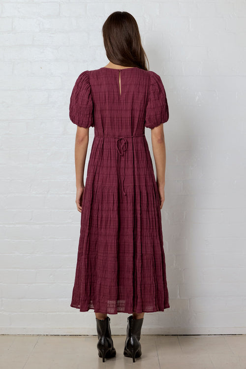Woman wearing a burgundy dress against a white brick wall.