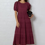 Woman wearing a burgundy dress with puff sleeves against a white brick wall.