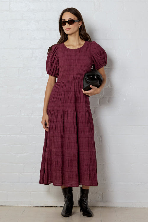 Woman wearing a burgundy dress with puff sleeves against a white brick wall.