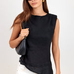 A woman modeling a sleeveless black top with white pants, black sunglasses, and a small black shoulder bag against a plain light background.