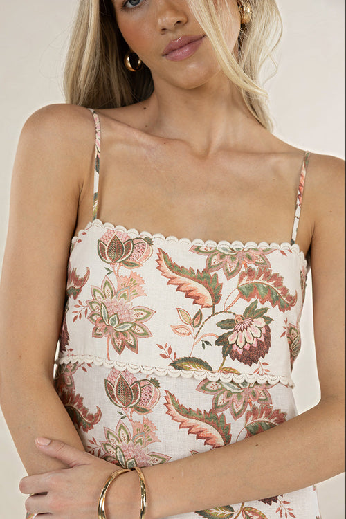 A close-up of a woman wearing a floral print camisole top with thin straps, a scalloped neckline, and a tiered ruffle detail at the waist. The top features a botanical pattern in muted pink, green, and brown tones on a cream background.