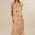 A woman modeling a sleeveless, floor-length maxi dress with an intricate orange, yellow, and beige floral mandala pattern against a warm beige studio backdrop.