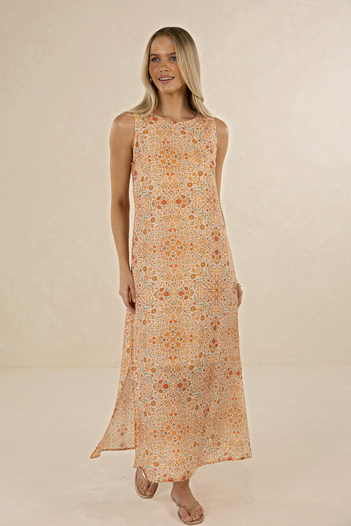 A woman modeling a sleeveless, floor-length maxi dress with an intricate orange, yellow, and beige floral mandala pattern against a warm beige studio backdrop.