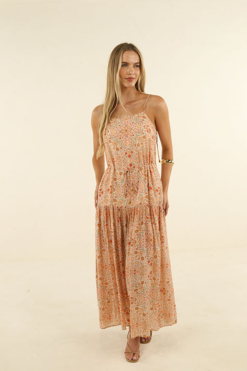 A woman modeling a sleeveless, tiered maxi dress with a small floral print in peach, orange, and cream tones, set against a plain beige studio background.