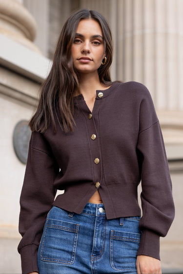 Woman wearing a brown cardigan and blue jeans standing in front of classical architecture.