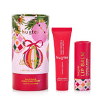 Huxter Lip & Hand Cream Duo Limited Edition set, featuring a cylindrical gift box with a pink and multicolor Christmas ornament design, a red lip balm tube, and a red lip balm tube with a matching patterned cap, on a white background.