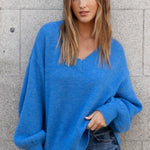 Woman wearing a blue sweater and jeans standing against a gray wall.