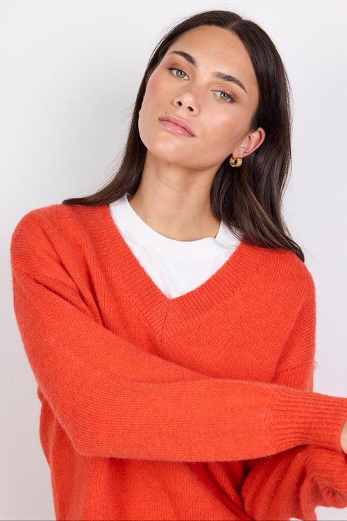 Woman wearing a orange sweater over a white shirt against a plain background