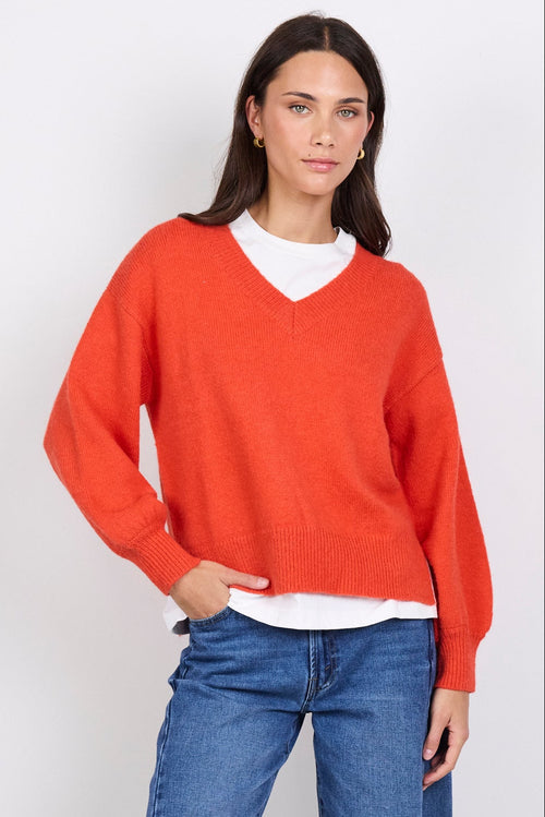 Person wearing an orange sweater, white shirt, blue jeans, and white sneakers on a white background