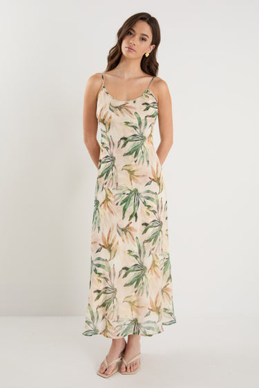Woman wearing a bias-cut midi dress with a tropical palm print, strappy sleeves, and a softly draped silhouette, styled with neutral sandals.