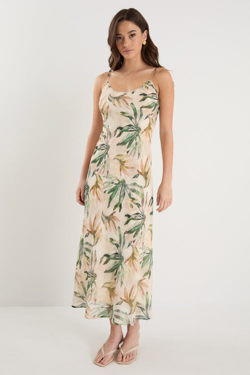 Woman wearing a bias-cut midi dress with a tropical palm print, strappy sleeves, and a softly draped silhouette, styled with neutral sandals.