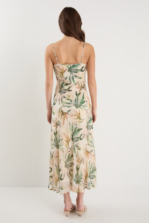 Woman wearing a bias-cut midi dress with a tropical palm print, strappy sleeves, and a softly draped silhouette, styled with neutral sandals.
