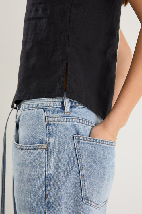 Woman wearing a sleeveless black linen top with contrast beige trim, styled with relaxed mid-blue denim jeans.