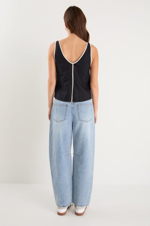 Woman wearing a sleeveless black linen top with contrast beige trim, styled with relaxed mid-blue denim jeans.