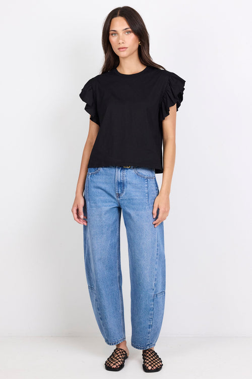 Woman wearing a black top with ruffles on the sleeves and blue jeans on a white background