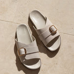 Pair of white sandals with gold buckles on a beige stone surface