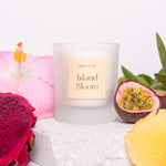 Candle labeled 'Island Bloom' surrounded by tropical fruits and flowers on a white surface.