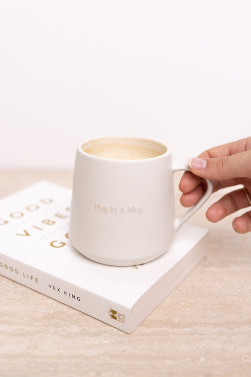Hand holding a white mug with 'Good Life' book on wooden surface