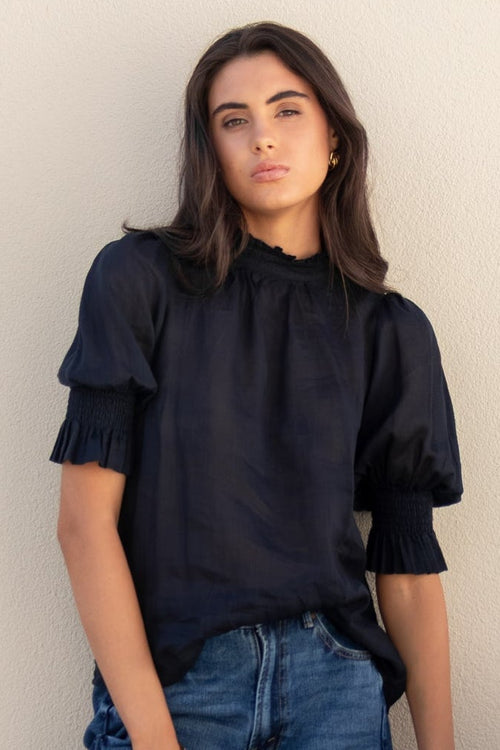 Woman in black top and blue jeans standing next to a large pot against a plain wall.