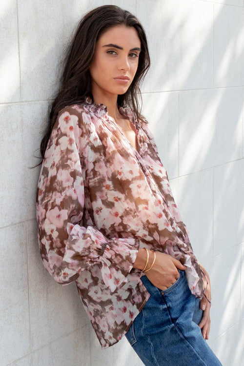 Woman leaning against a white wall wearing a floral blouse and blue jeans.