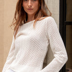 Woman wearing a textured white sweater and white pants against a beige wall.