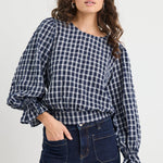 Woman wearing a blue and white checkered blouse with puffed sleeves and dark jeans on a white background