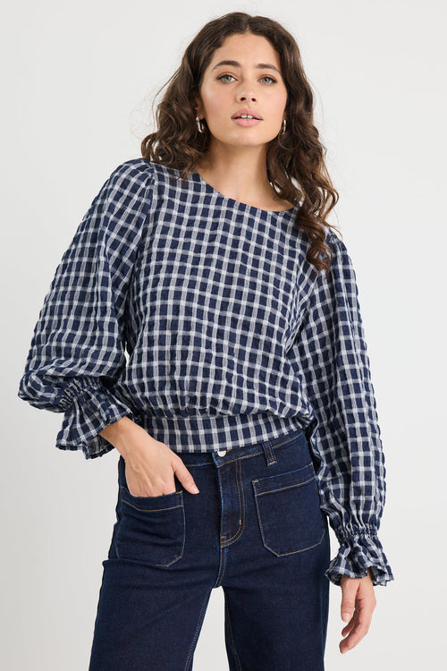 Woman wearing a blue and white checkered blouse with puffed sleeves and dark jeans on a white background