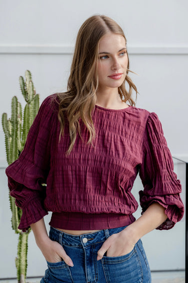 Woman wearing a burgundy long sleeve top with shirred detailing and fluted cuffs, styled with high-waisted blue jeans.