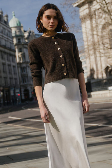 Woman in a brown cardigan and white skirt standing on a city street with buildings in the background