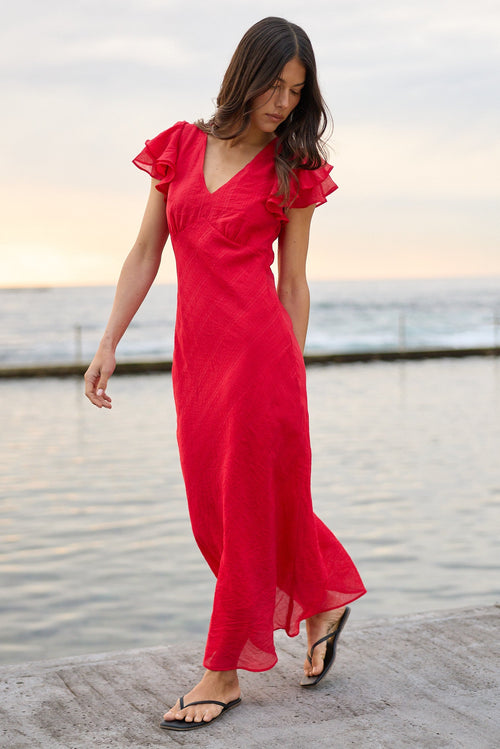 A woman wearing a bright red maxi dress with ruffled short sleeves walks along a concrete pier next to a calm body of water under an overcast sky.