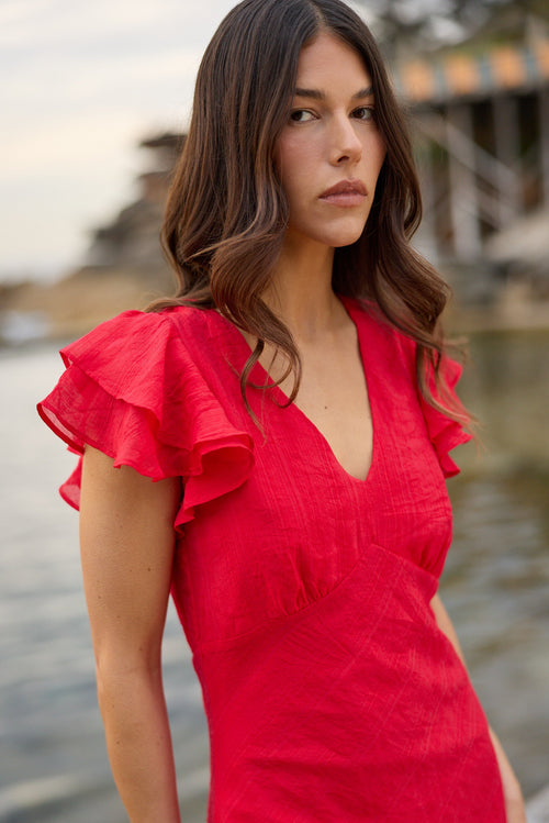 A woman wearing a bright red wrap-style dress with ruffled short sleeves, standing outdoors near water with a blurred waterfront setting in the background.