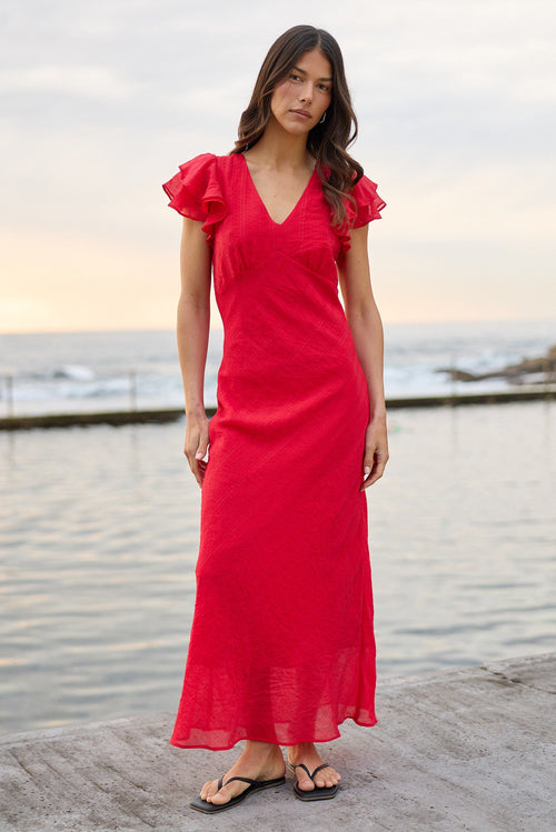 A woman wearing a bright red maxi dress with ruffled short sleeves and a V-neckline stands on a concrete pier with the ocean and cloudy sky in the background.