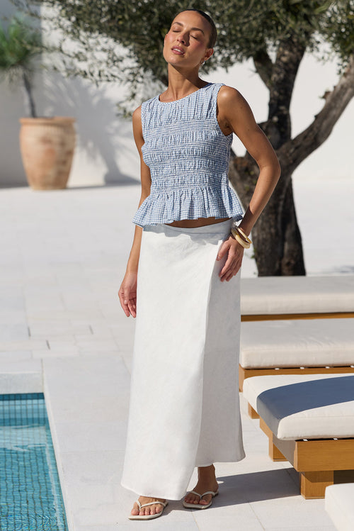 A woman stands by a poolside patio wearing a light blue smocked sleeveless top and a long white skirt. White lounge chairs, a tree, and a tiled pool are visible in the background.