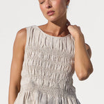 A woman wearing a sleeveless, smocked beige top and blue jeans, standing against a bright white studio background.
