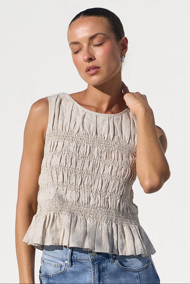 A woman wearing a sleeveless, smocked beige top and blue jeans, standing against a bright white studio background.