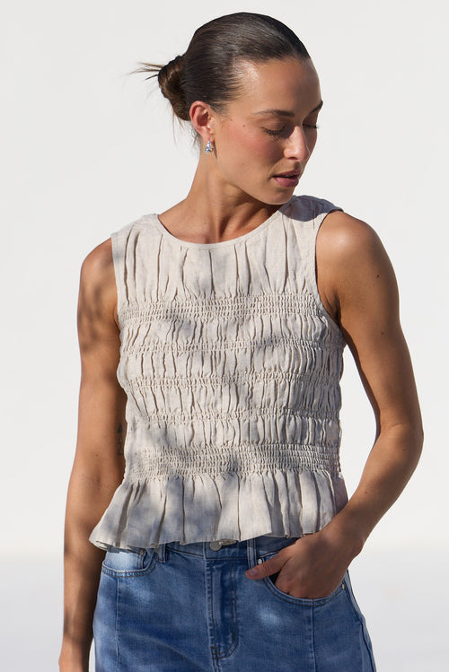 A woman wearing a sleeveless, smocked beige top and blue jeans, standing against a bright white studio background.