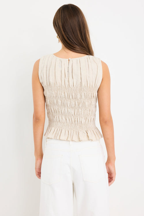 Woman wearing a natural linen sleeveless top with shirred bodice and ruffle hem, styled with ecru jeans.