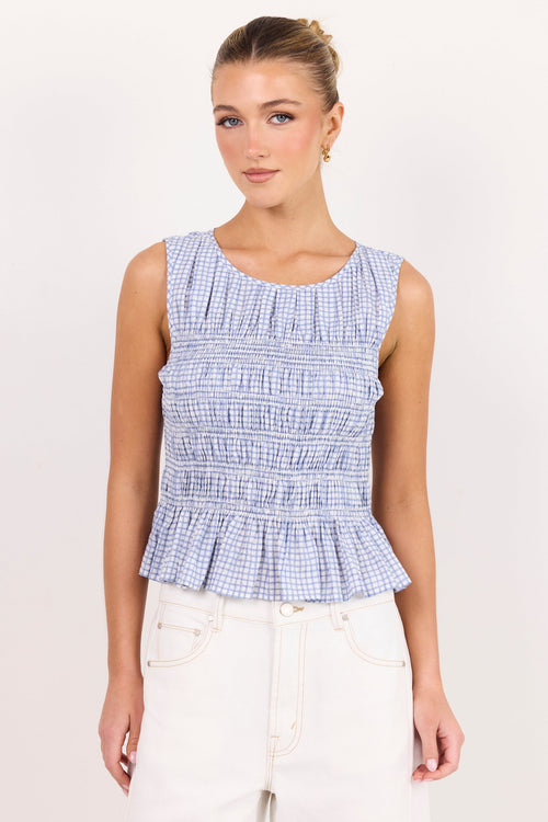 A woman modeling a sleeveless smocked top with a light blue and white gingham pattern, paired with white pants, against a plain off-white studio background.