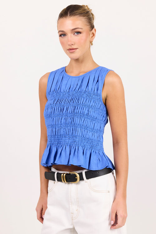 A woman modeling a bright blue sleeveless smocked top with a ruffled hem, paired with white pants and a black belt, against a plain white studio background.