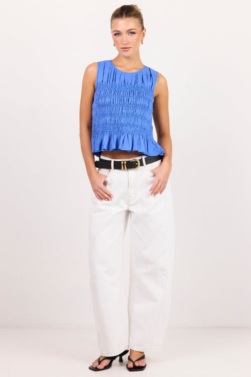 A woman modeling a sleeveless smocked blue top with a ruffled hem, paired with white wide-leg jeans, a black belt, and black sandals, against a plain white studio background.