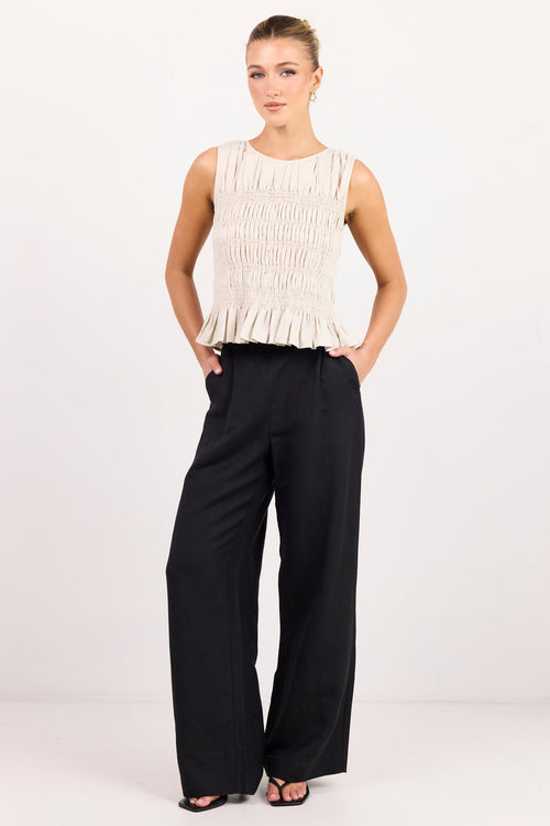 A woman modeling a sleeveless cream pleated top and black wide-leg trousers against a plain white studio background.