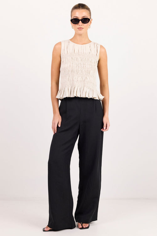A model stands against a plain light gray studio background wearing a sleeveless cream pleated top with a ruffled hem and black wide-leg trousers.