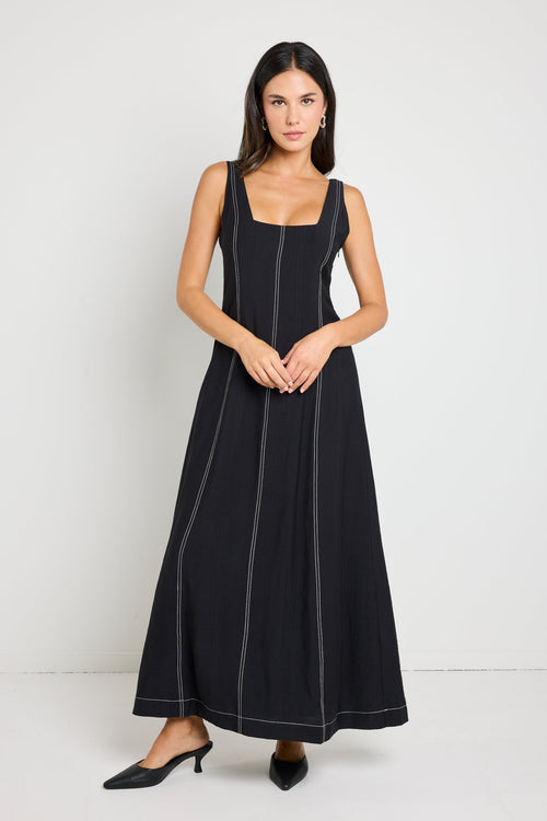 Woman wearing a black maxi dress with a square neckline and contrast stitching, styled with black heels for a chic, modern look.