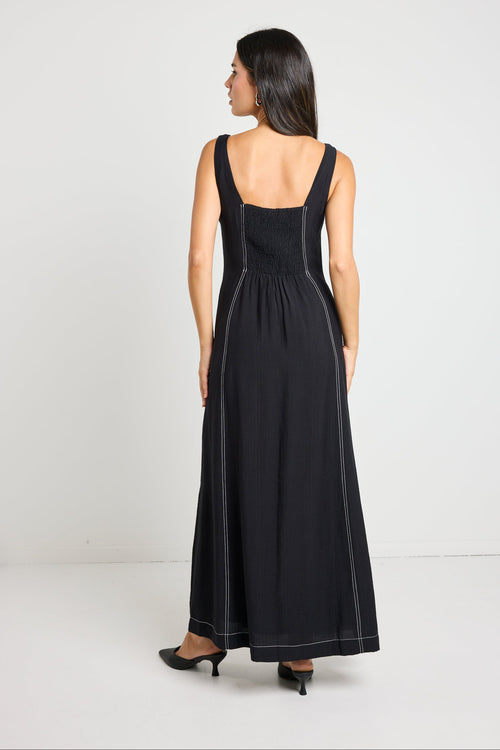 Woman wearing a black maxi dress with a square neckline and contrast stitching, styled with black heels for a chic, modern look.